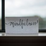 The word mindfulness written in cursive in black ink on white paper rests on a windowsill. The window has black panes and the sky appears cloudy outside.